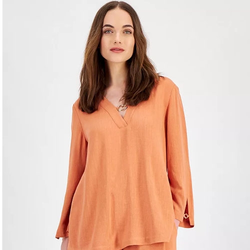 a woman with brown hair wearing a orange colored long sleeve top in front of a light grey background