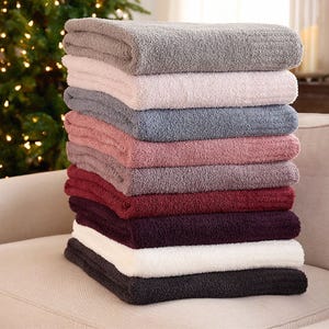 A stack of plush, soft Barefoot Dreams CozyChic blankets in various colors, including shades of gray, pink, burgundy, and cream, is arranged on a beige sofa, with a Christmas tree in the background.