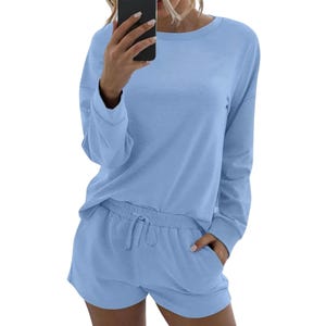 A person wearing a light blue loungewear set consisting of a long-sleeve top and drawstring shorts, taking a mirror selfie with a smartphone.