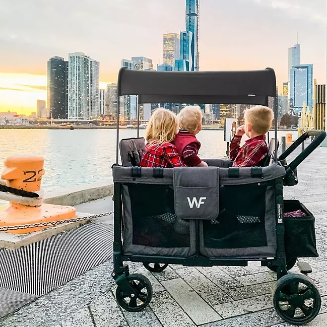 Three children are seated in a large black wagon stroller with a canopy, facing a city waterfront.