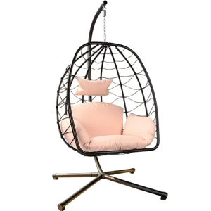 A hanging egg chair with a metal frame and light pink cushions is situated on a patio, surrounded by greenery and palm trees.