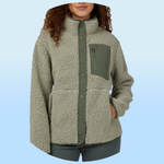 A woman wearing a light green, button-up sherpa fleece jacket with a high collar, featuring a contrasting green chest pocket with a zipper and side pockets.