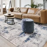 A modern living room setup featuring a tan leather couch, a plush blue ottoman, and two round nesting coffee tables on a gray and blue abstract-patterned area rug.