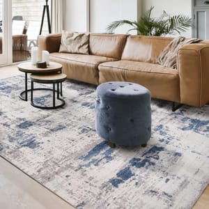 A modern living room setup featuring a tan leather couch, a plush blue ottoman, and two round nesting coffee tables on a gray and blue abstract-patterned area rug.