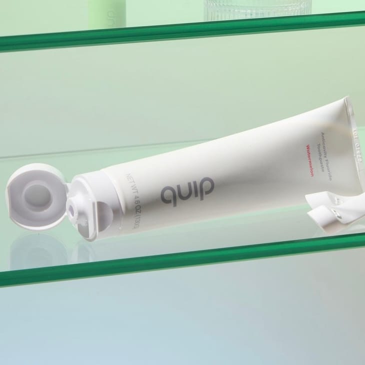 A tube of quip toothpaste with an open cap, labeled \“Watermelon,\“ resting on a glass shelf.