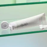 A tube of quip toothpaste with an open cap, labeled \“Watermelon,\“ resting on a glass shelf.