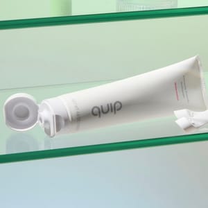 A tube of quip toothpaste with an open cap, labeled \“Watermelon,\“ resting on a glass shelf.