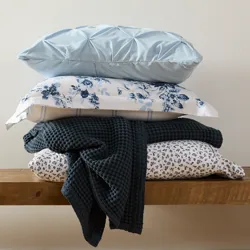A stack of pillows and a blanket are displayed on a wooden bench. The pillows feature various designs, including solid light blue, floral, and checkered patterns, complemented by a textured, dark-colored blanket.