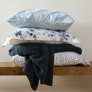 A stack of pillows and a blanket are displayed on a wooden bench. The pillows feature various designs, including solid light blue, floral, and checkered patterns, complemented by a textured, dark-colored blanket.