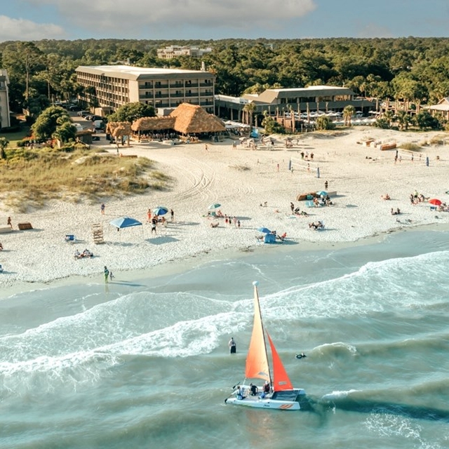 A sailboat with an orange sail navigates near the shore of a beach with people and umbrellas.