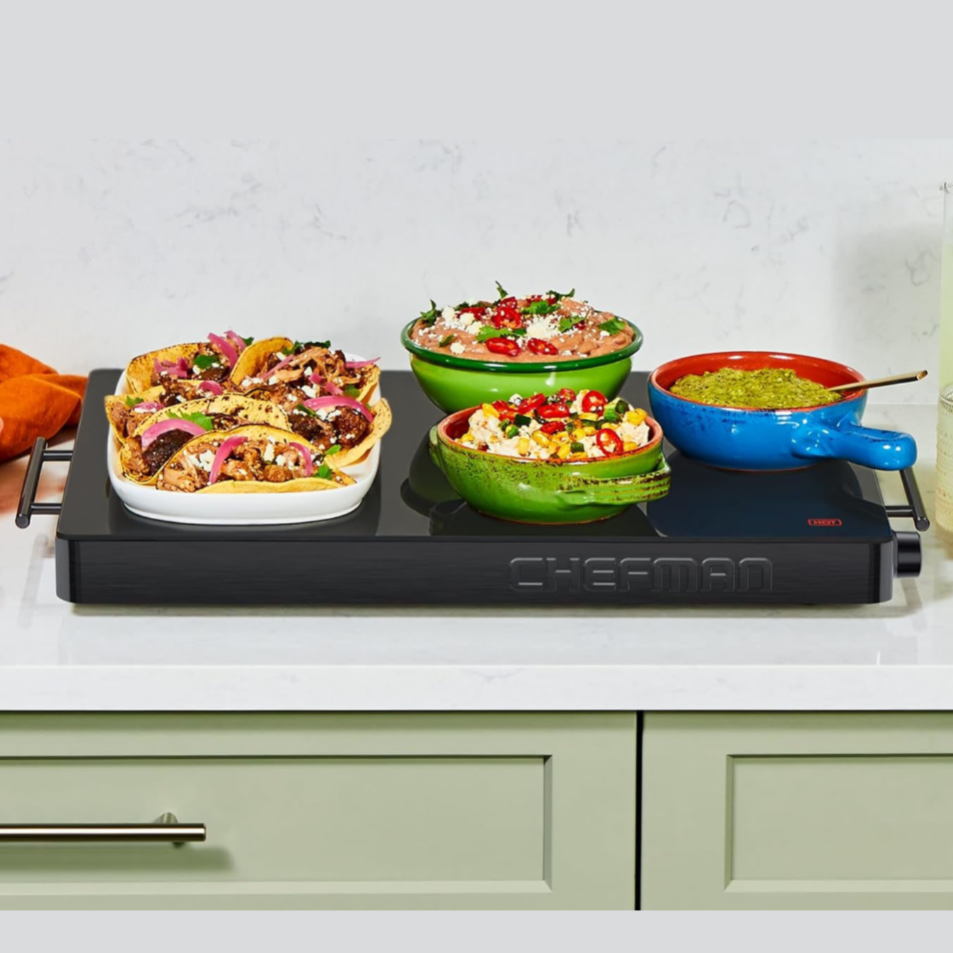 A Chefman warming tray holds various dishes, including nachos and dips, in colorful bowls.