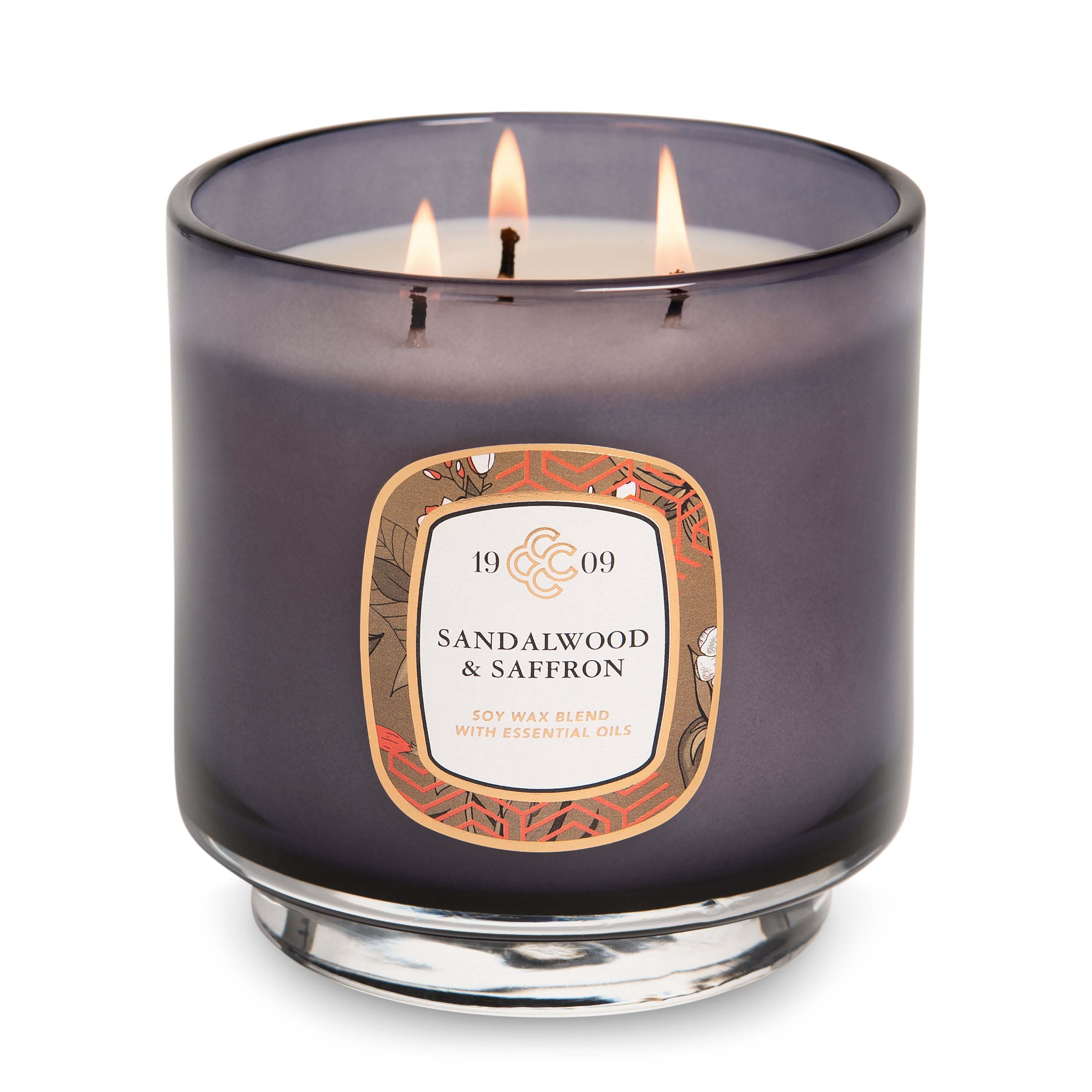 A sandalwood and saffron scented soy wax blend candle with essential oils, featuring a dark glass container and three lit wicks.