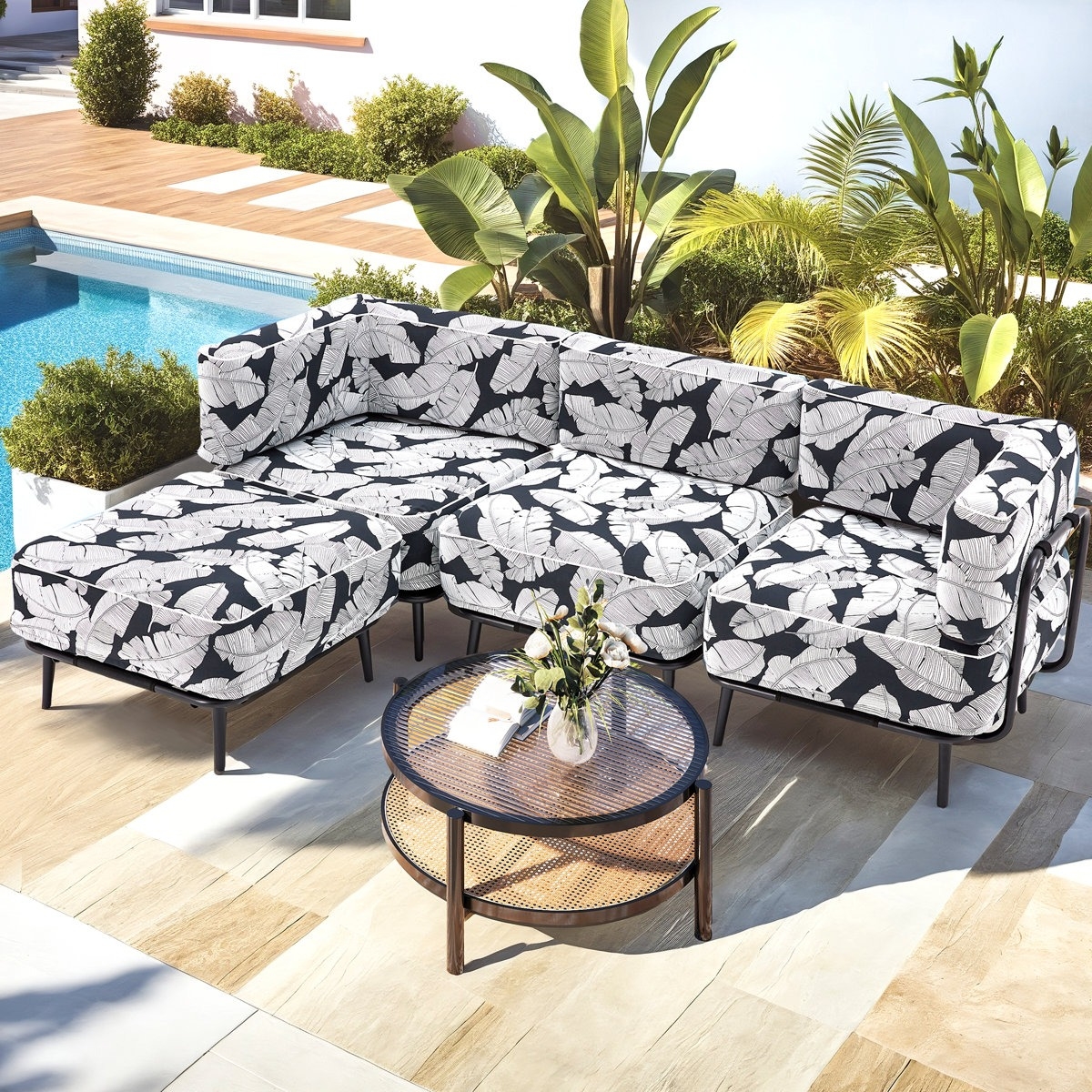 Outdoor sectional sofa with black and white leaf-patterned cushions next to a round wicker and glass coffee table, set on a patio by a pool with surrounding plants and greenery.