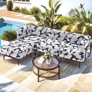 Outdoor sectional sofa with black and white leaf-patterned cushions next to a round wicker and glass coffee table, set on a patio by a pool with surrounding plants and greenery.