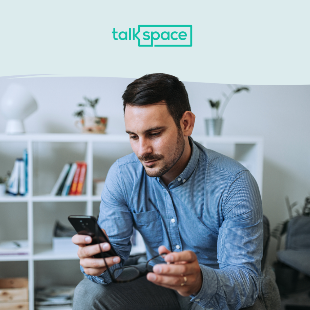 A person wearing a blue shirt holds a smartphone and glasses, with a Talkspace logo above. In the background is a bookshelf with books and plants.