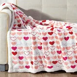 A plush blanket with a heart pattern in various shades of pink and red, featuring the phrase \“be mine,\“ draped over a beige sofa.