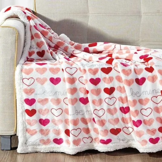 A plush blanket with a heart pattern in various shades of pink and red, featuring the phrase \“be mine,\“ draped over a beige sofa.