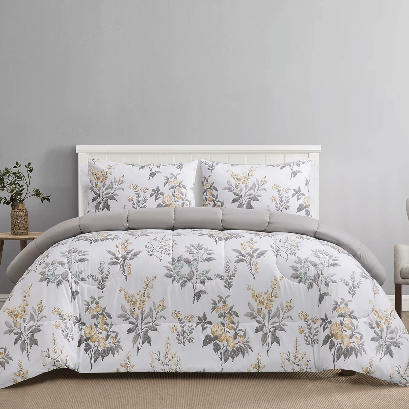 A bed set with a floral-patterned comforter and matching pillowcases in shades of grey, yellow, and white, accessorized with a small wooden side table holding a potted plant.
