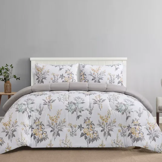 A bed set with a floral-patterned comforter and matching pillowcases in shades of grey, yellow, and white, accessorized with a small wooden side table holding a potted plant.