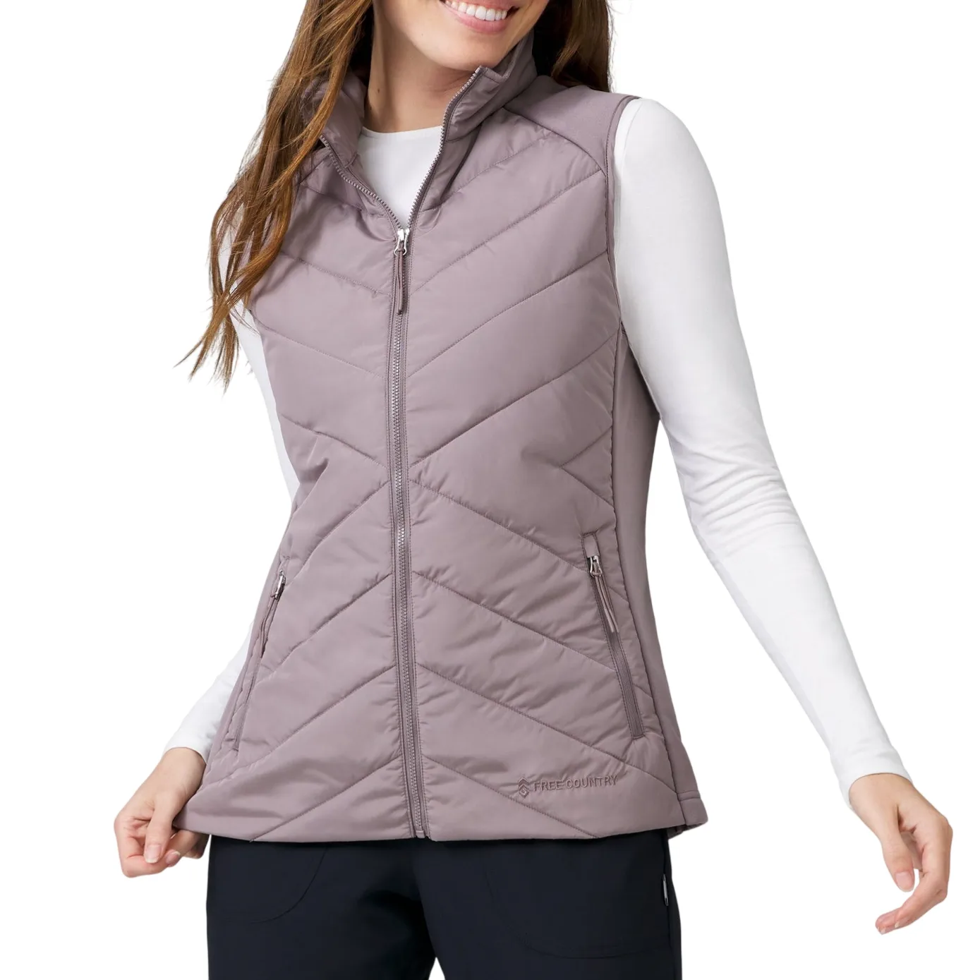 A woman wears a light mauve quilted vest with a high collar and two zippered pockets, paired with a white long-sleeve shirt and black pants.