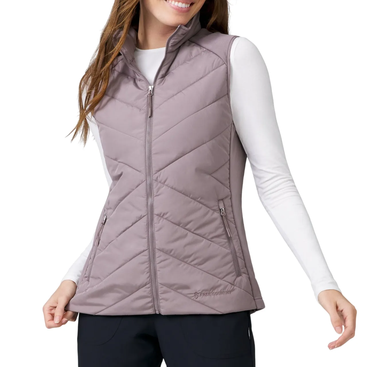 A woman wears a light mauve quilted vest with a high collar and two zippered pockets, paired with a white long-sleeve shirt and black pants.