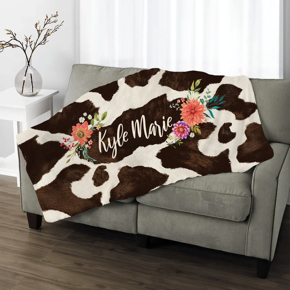 Cowhide-patterned throw blanket with floral accents and the name \“Kyle Marie\“ embroidered, draped over a gray couch.