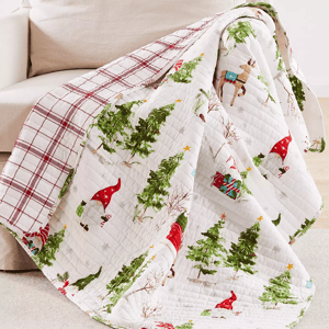 A quilt with a holiday theme featuring Christmas trees, reindeer, gifts, and Santa hats draped over a couch.