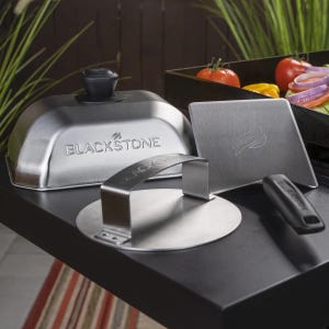 Blackstone stainless steel griddle accessories including a basting dome, press, and scraper, with a variety of vegetables being cooked on a griddle in the background.