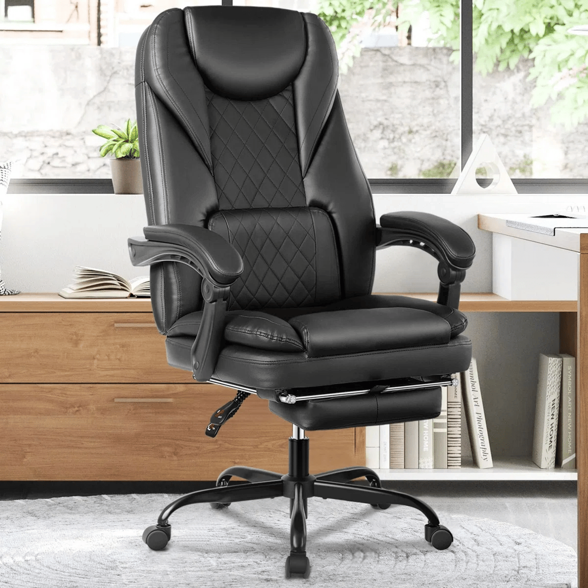 A black leather office chair with a high back, quilted design, adjustable armrests, and a retractable footrest, placed on a gray carpet in front of a wooden cabinet and window.