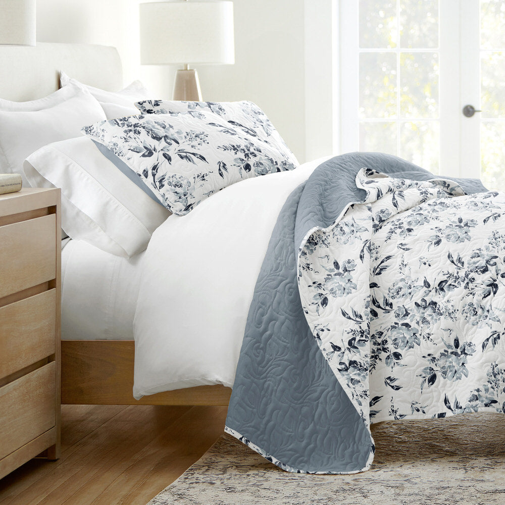 A bed with a blue and white floral comforter, matching pillowcases, and a light gray quilt is shown. A wooden nightstand, table lamp, and large window with sheer curtains complete the setup.