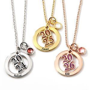 The 2025 graduation necklace features a gold tone circular pendant with \“2025\“ in the center, a white gemstone, and the name \“Emma\“ engraved. It hangs on a matching chain.