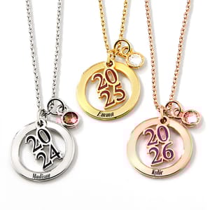 The 2025 graduation necklace features a gold tone circular pendant with \“2025\“ in the center, a white gemstone, and the name \“Emma\“ engraved. It hangs on a matching chain.