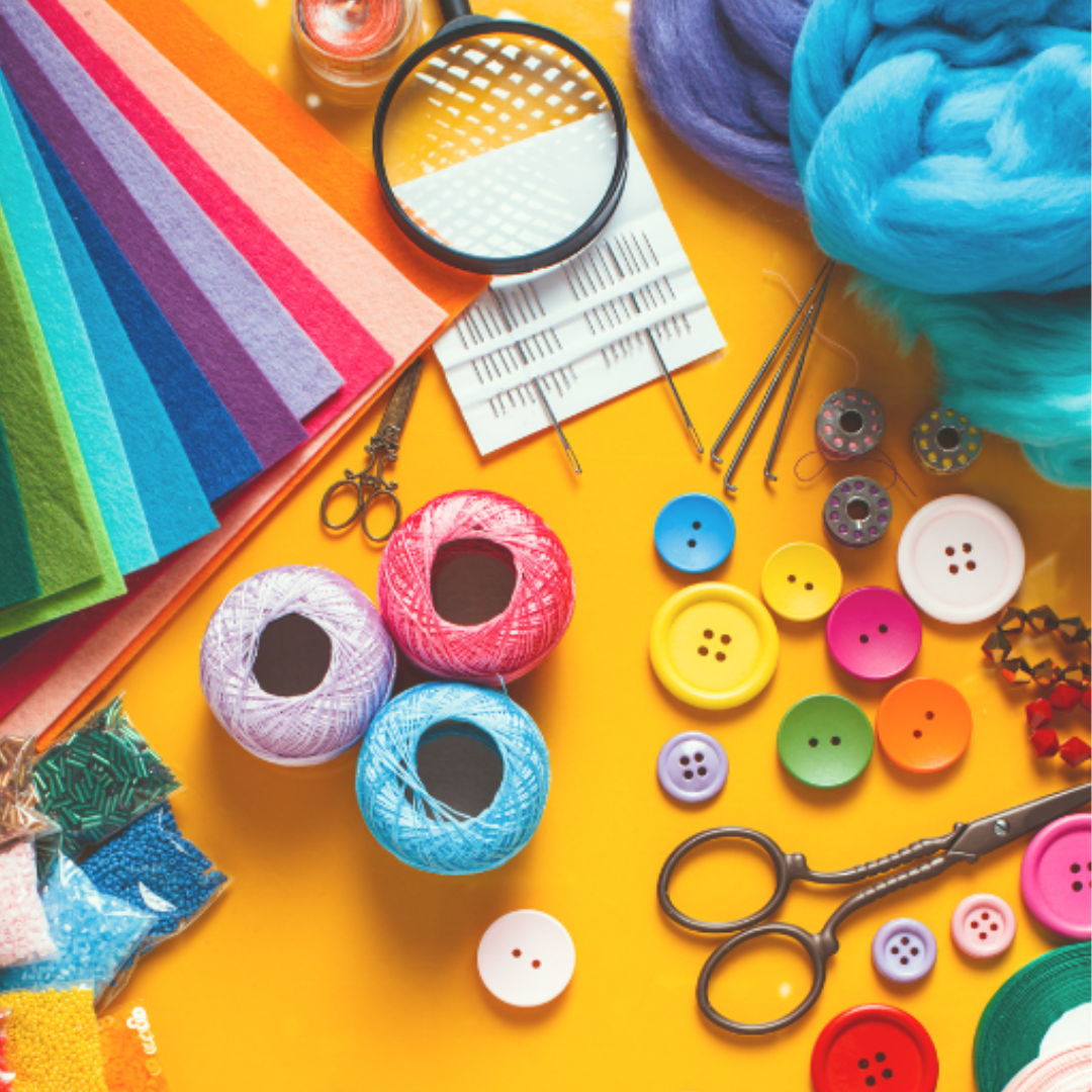 Craft supplies including colorful felt sheets, yarn, buttons, scissors, sewing needles, crochet hooks, thread bobbins, beads, and a magnifying glass are arranged on a yellow surface.