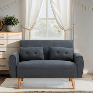 A dark gray loveseat with wooden legs is centered in a room, accompanied by two matching cushions. A light-colored dresser with brass handles and a potted plant is positioned on the left, and sheer white curtains hang from a window in the background.