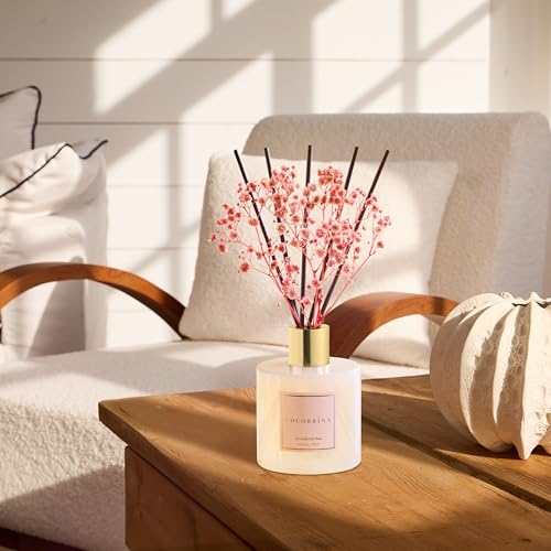 A room diffuser with pink flowers and reeds is placed on a wooden table beside a white textured vase in a sunlit, cozy seating area with wooden armchairs.