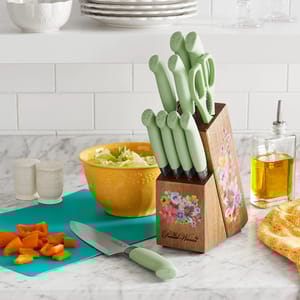 A set of kitchen knives with pastel green handles in a wooden block, a yellow mixing bowl with salad, and some sliced carrots on a teal cutting board.