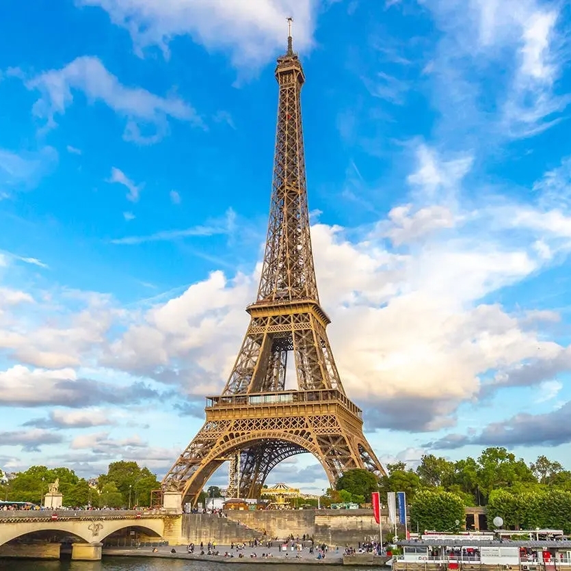 Eiffel Tower with blue sky and clouds in the background, a bridge, and people gathered near the base.