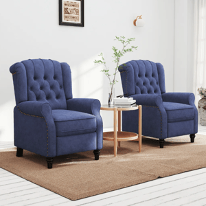 Two blue, tufted recliner chairs with nailhead trim are arranged on a beige rug, separated by a small round wooden table holding a plant and books. White walls with sconce lighting and a framed artwork in the background complete the cozy setting.