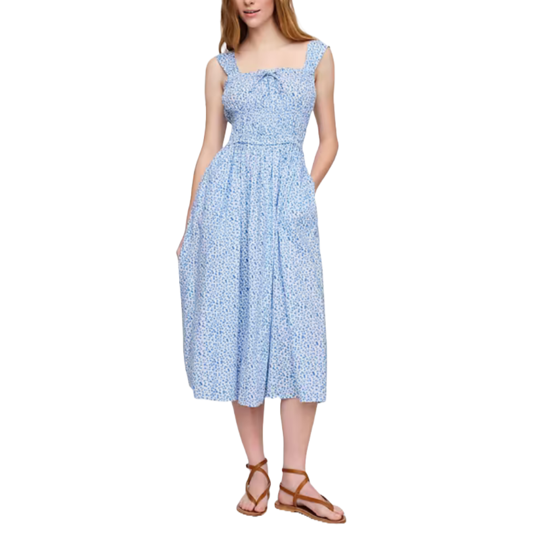 A blue smocked midi dress with a subtle floral pattern, featuring wide straps and a gathered bodice. The dress has a full skirt with pockets and is paired with brown strappy flat sandals.