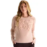 A woman wearing a light pink sweater featuring textured flower designs on the chest area, paired with dark trousers.
