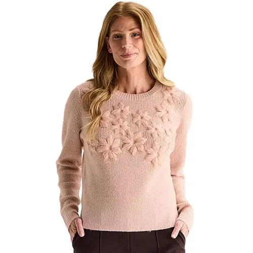 A woman wearing a light pink sweater featuring textured flower designs on the chest area, paired with dark trousers.