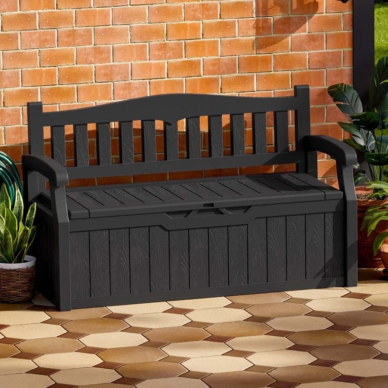 A black outdoor bench with a storage compartment is set against a brick wall, surrounded by potted plants, on a tiled patio.