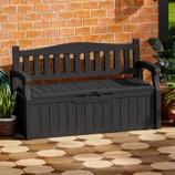 A black outdoor bench with a storage compartment is set against a brick wall, surrounded by potted plants, on a tiled patio.