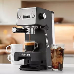 Cuisinart espresso maker with a portafilter and a steam wand, accompanied by a glass of iced coffee and two stacked white mugs in a kitchen setting.