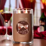 Yankee Candle labeled \“Cozy Cocktail Lounge,\“ scented with sweet vermouth and vetiver, features two lit wicks. A cocktail lounge setting with a drink and a wine bottle is visible in the background.