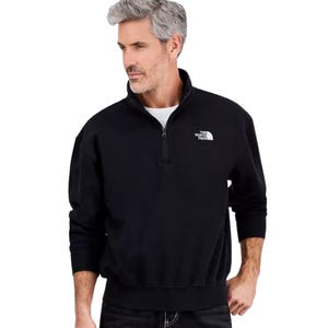 A man wears a black North Face quarter-zip sweatshirt with a high collar and the brand logo on the left chest. The sweatshirt has ribbed cuffs and hem, offering a casual look.