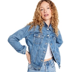 A woman is wearing a denim jacket with button closures over a white top, paired with denim jeans. She has curly hair and a casual pose.