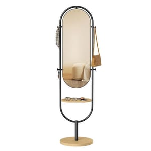 A modern, oval full-length floor mirror with a black metal frame and a wooden base, featuring side hooks for hanging items and a small circular wooden shelf below for accessories.