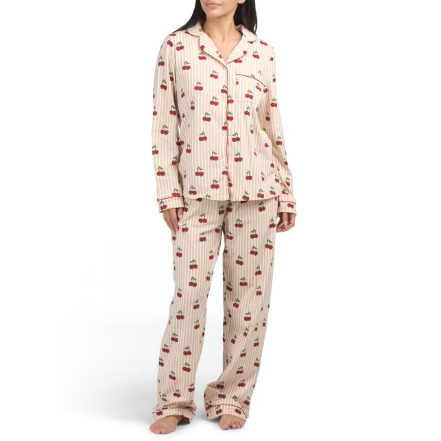 A woman is wearing a pajama set featuring a beige color with cherry patterns. The set includes a long-sleeved button-up top and matching pants with vertical stripes.