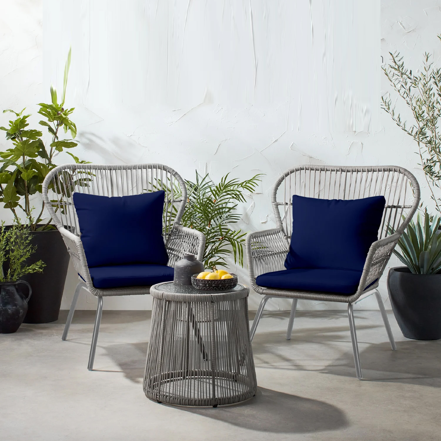 Two grey wicker chairs with blue cushions, accompanied by a grey round wicker table, surrounded by potted plants.